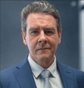 Portrait of a middle-aged man in a blue suit and light gray tie against a blurred blue background, facing the camera. It appears to be a formal headshot for business use.